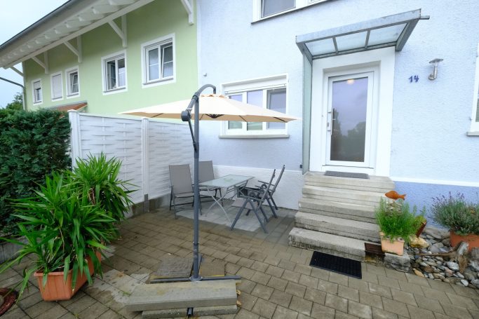 Eingangsbereich mit Terrasse, Tisch, Stühlen und Sonnenschirm vor einem Wohnhaus.